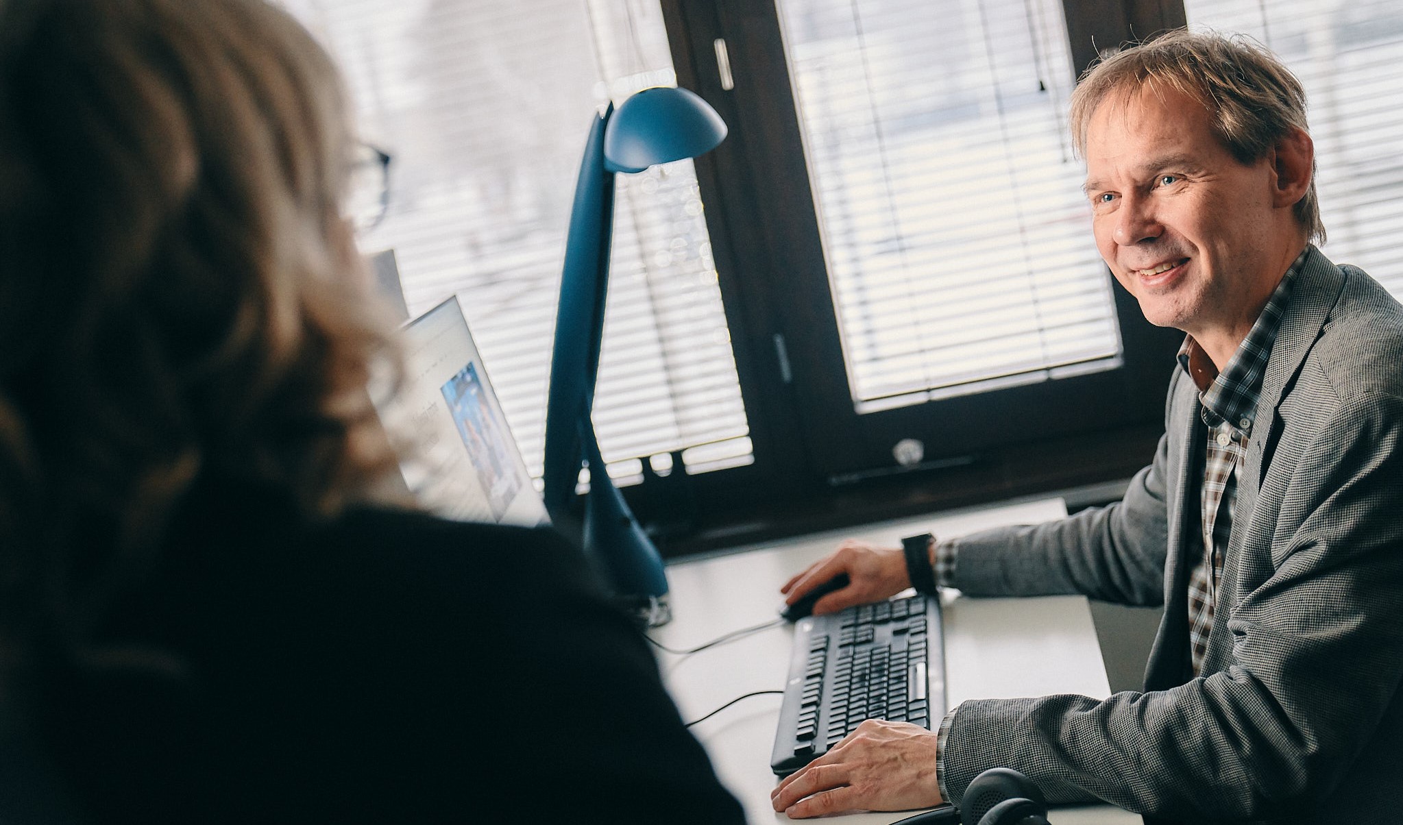 Two persons in an office. One person's back is facing the camera. The other person has a smile on his face.