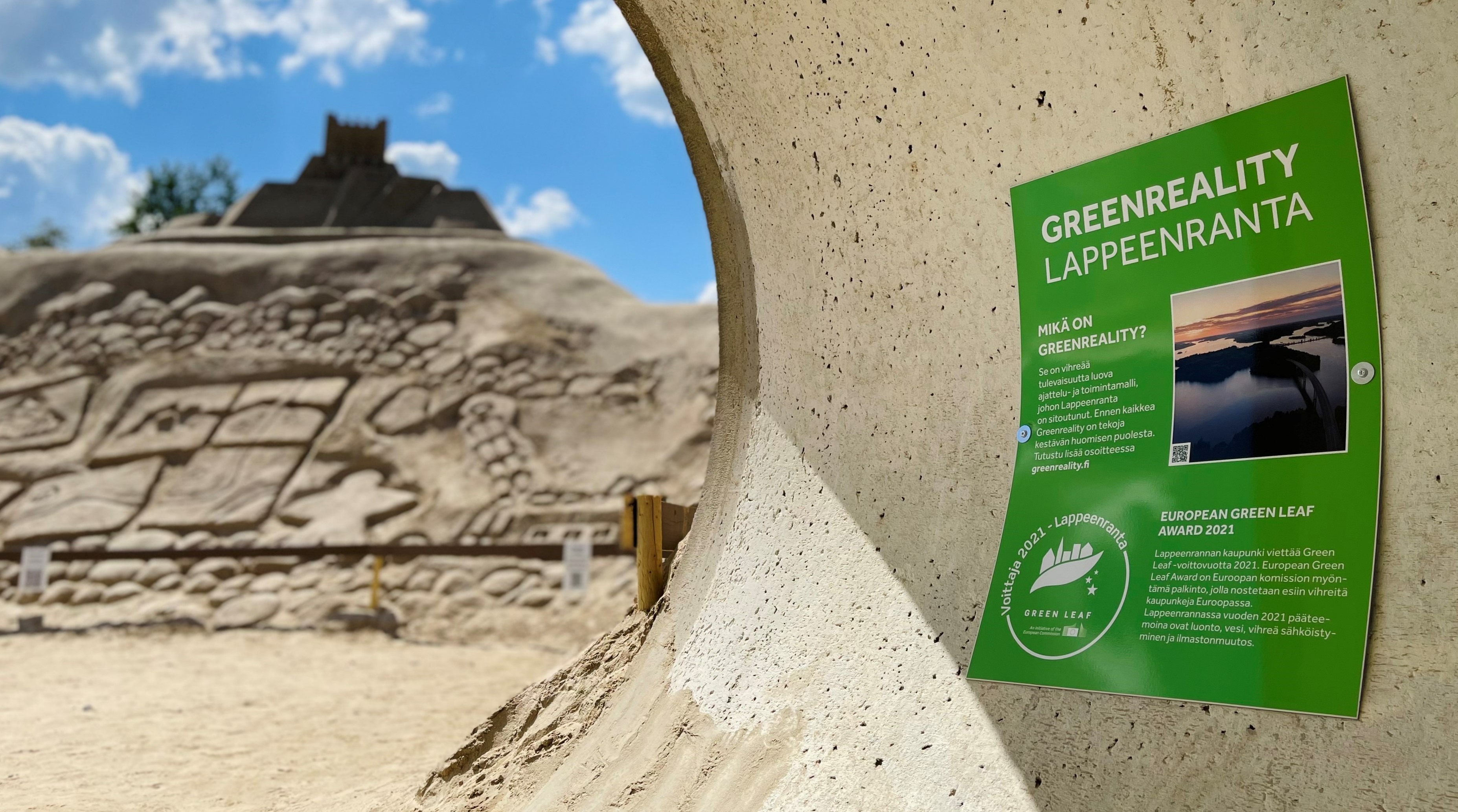 A green info sign with Greenreality Lappeenranta written with big letters. Blurry in the background, sculptures of the sandcastle and a blue sky.