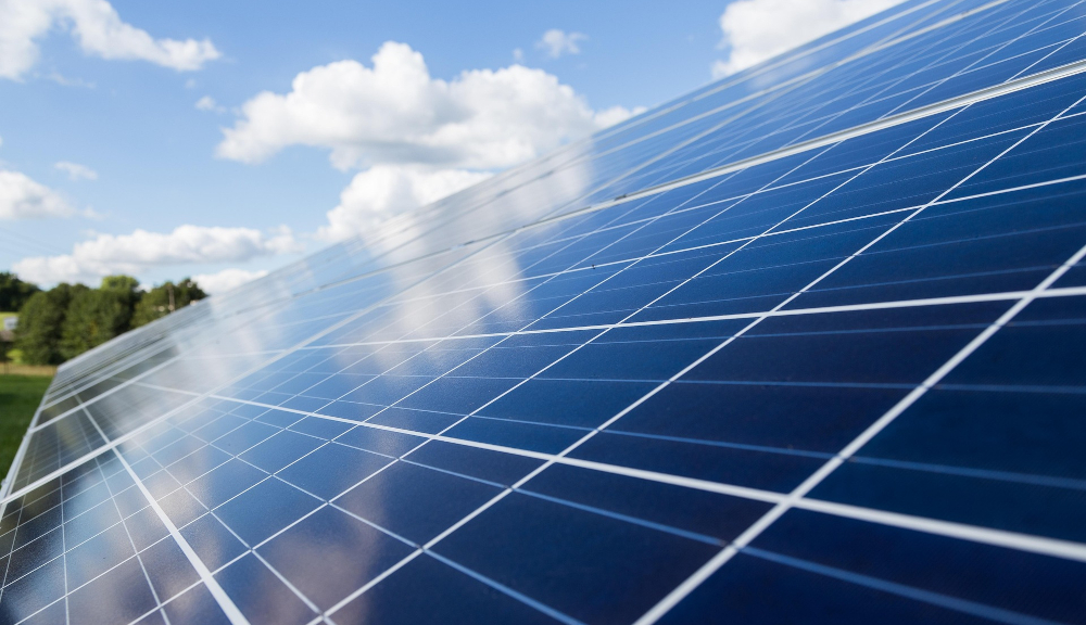 A solar panel with a background of blue sky, clouds, and a forest edge.